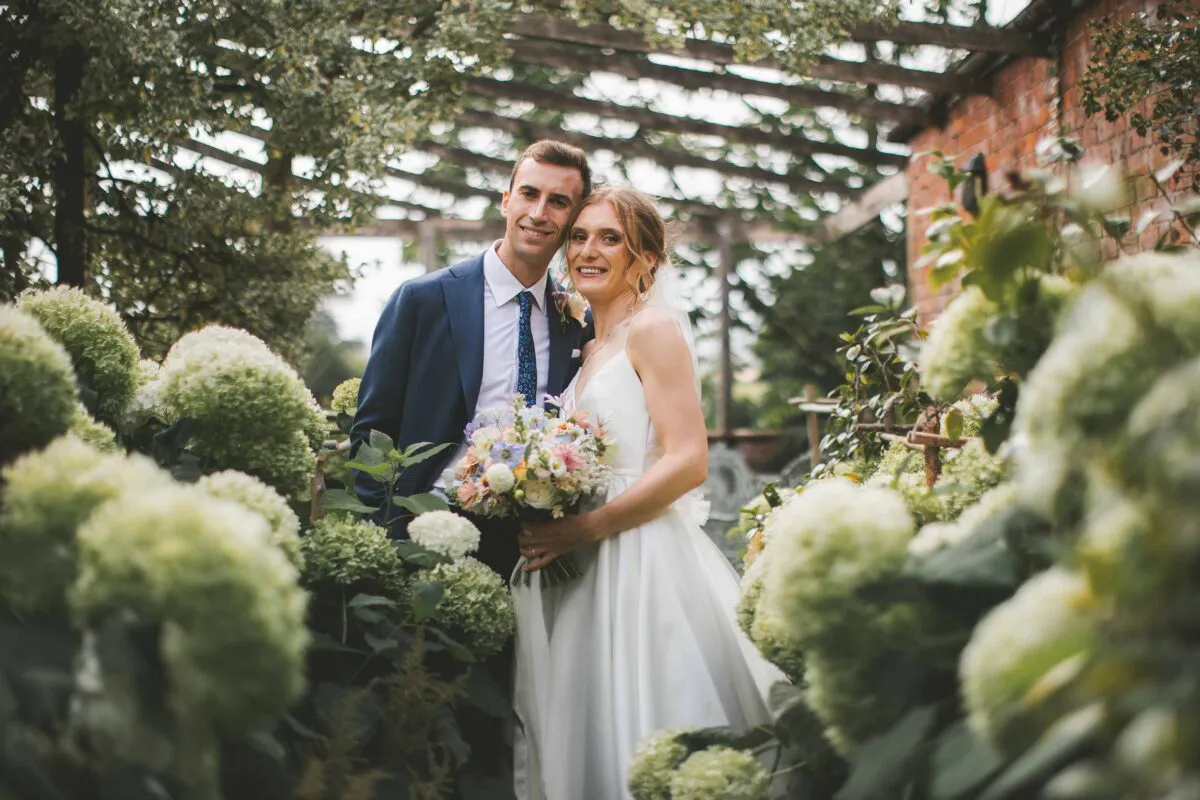A smiling bride and groom stand together surrounded by lush green hydrangea bushes and an overhead pergola structure in a gorgeous garden setting.
