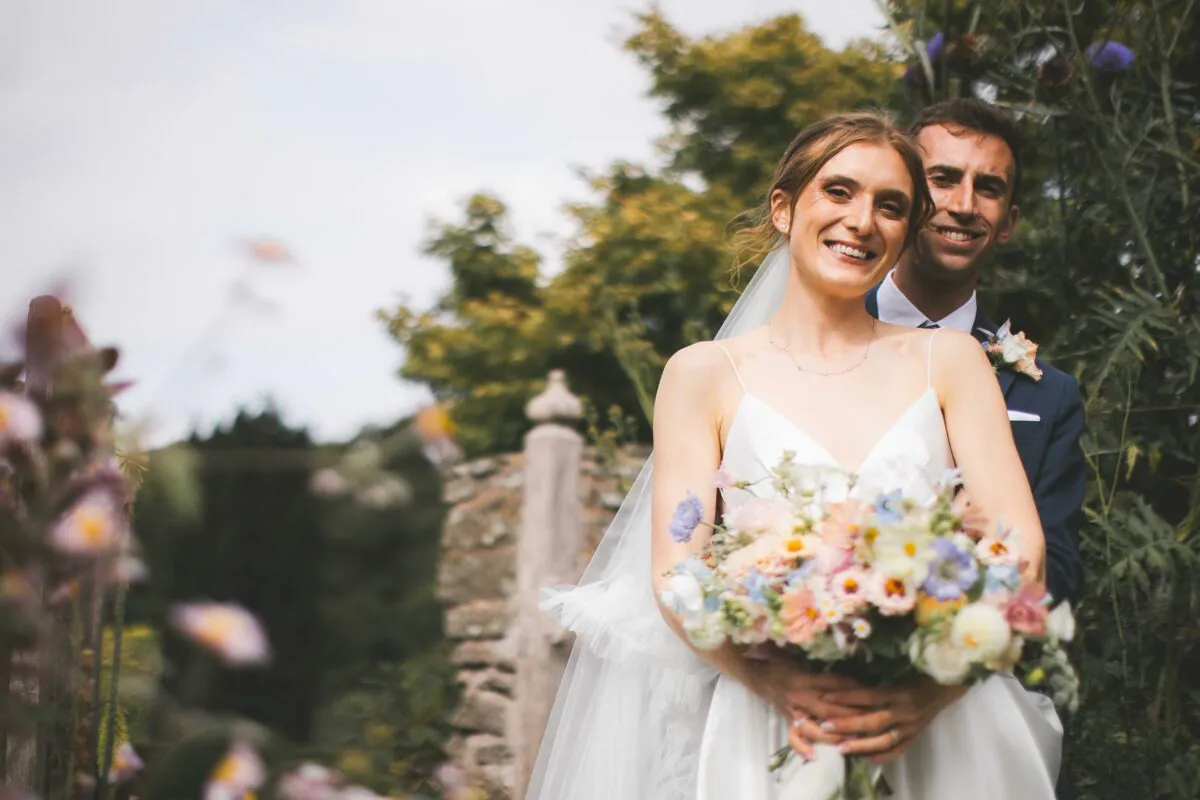 A smiling newlywed couple embraces outdoors in a lush garden setting, with the bride holding a colourful pastel bouquet and wearing a sleek slip dress and veil.