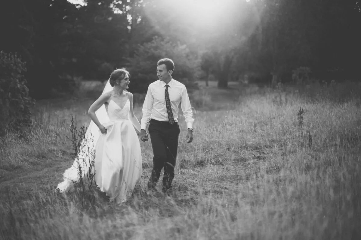 A newlywed couple walk hand in hand through a tall grass field while sharing a joyful glance under soft backlit sunlight.
