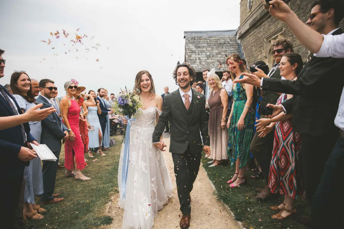 A joyful newlywed couple walks hand in hand down an outdoor path while smiling guests shower them in colourful confetti outside a stone church.