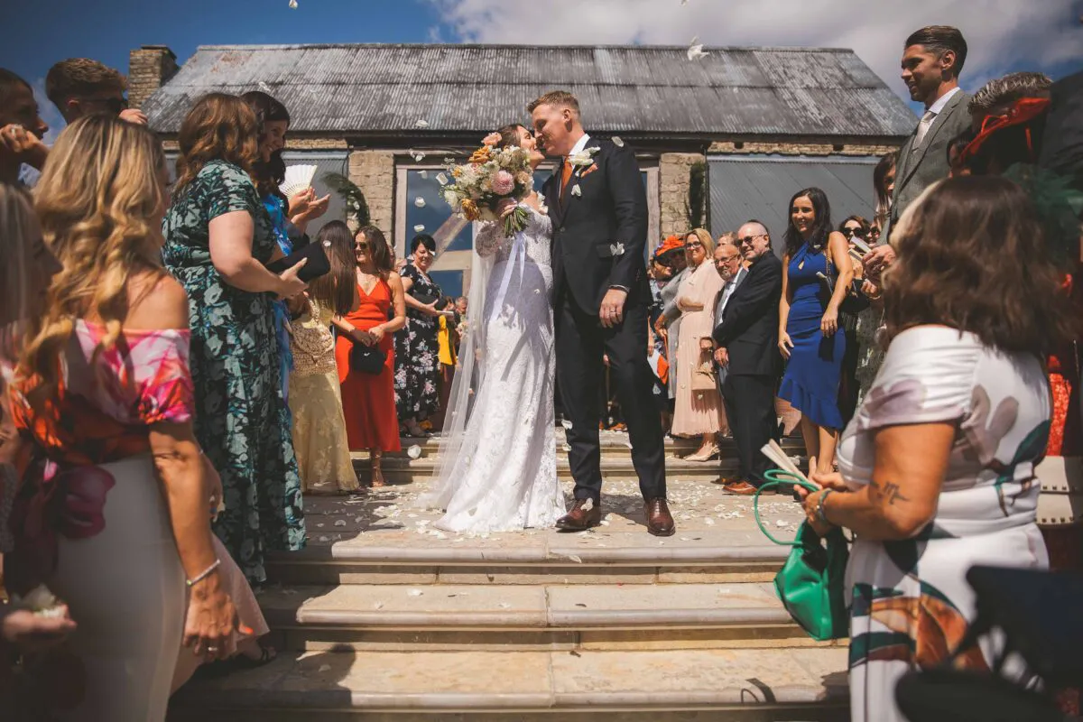 The newlywed couple kiss as guests shower them with confetti on stone steps outside a rustic stone barn on a sunny wedding day.
