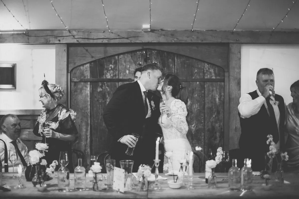 A newlywed couple share a kiss at the top table during wedding speeches in a rustic barn venue decorated with twinkling fairy lights and candlelit tables.