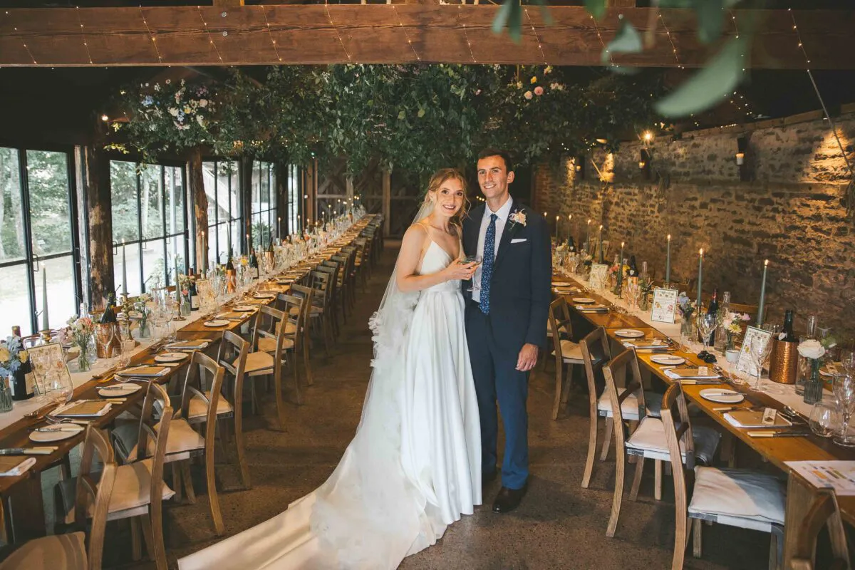 A newlywed couple smiles together in a rustic barn wedding venue featuring long wooden banquet tables, exposed brick walls, and a suspended floral ceiling installation.