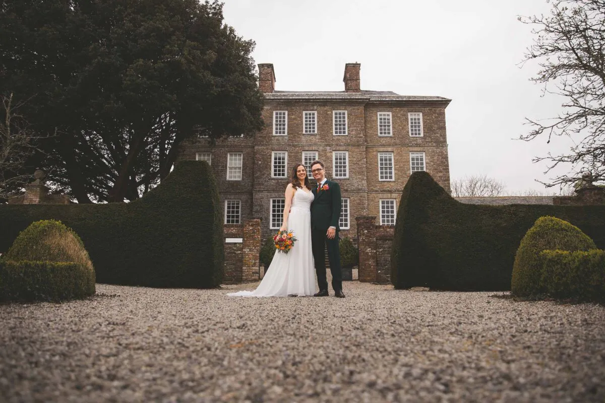 Newlyweds pose together on a gravel path in front of a grand stone manor house framed by manicured hedges and large trees.