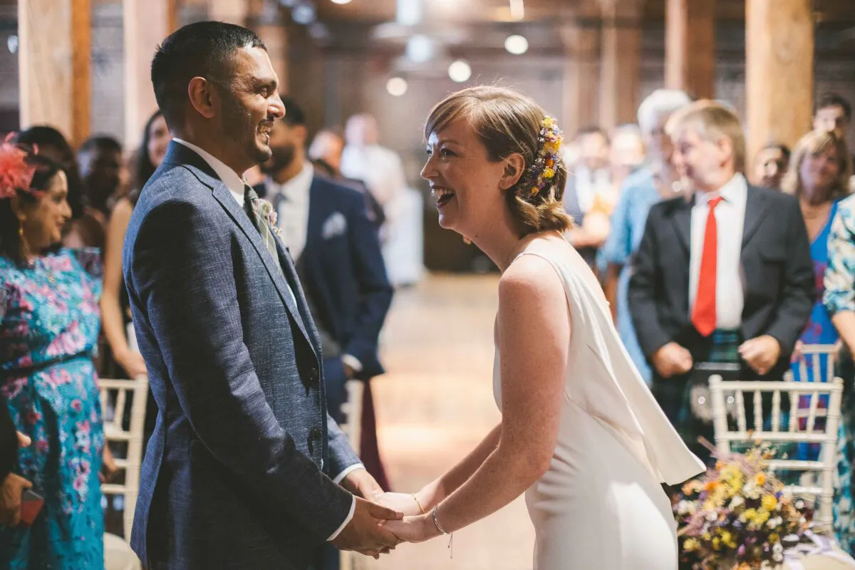 A joyful bride and groom hold hands and share a laugh during their wedding ceremony in a rustic barn venue with guests looking on.