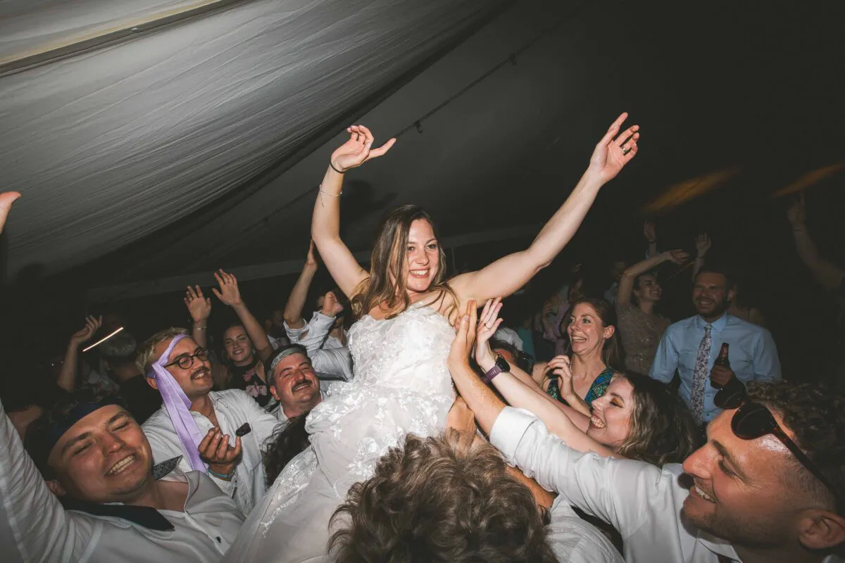 A joyful bride in a strapless lace wedding dress is lifted high into the air by a cheering crowd of wedding guests on a lively dance floor.