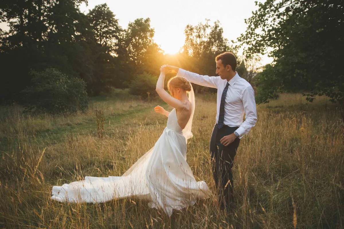 Newlyweds share a romantic dance together in a sunlit meadow during golden hour, surrounded by tall grass and trees.