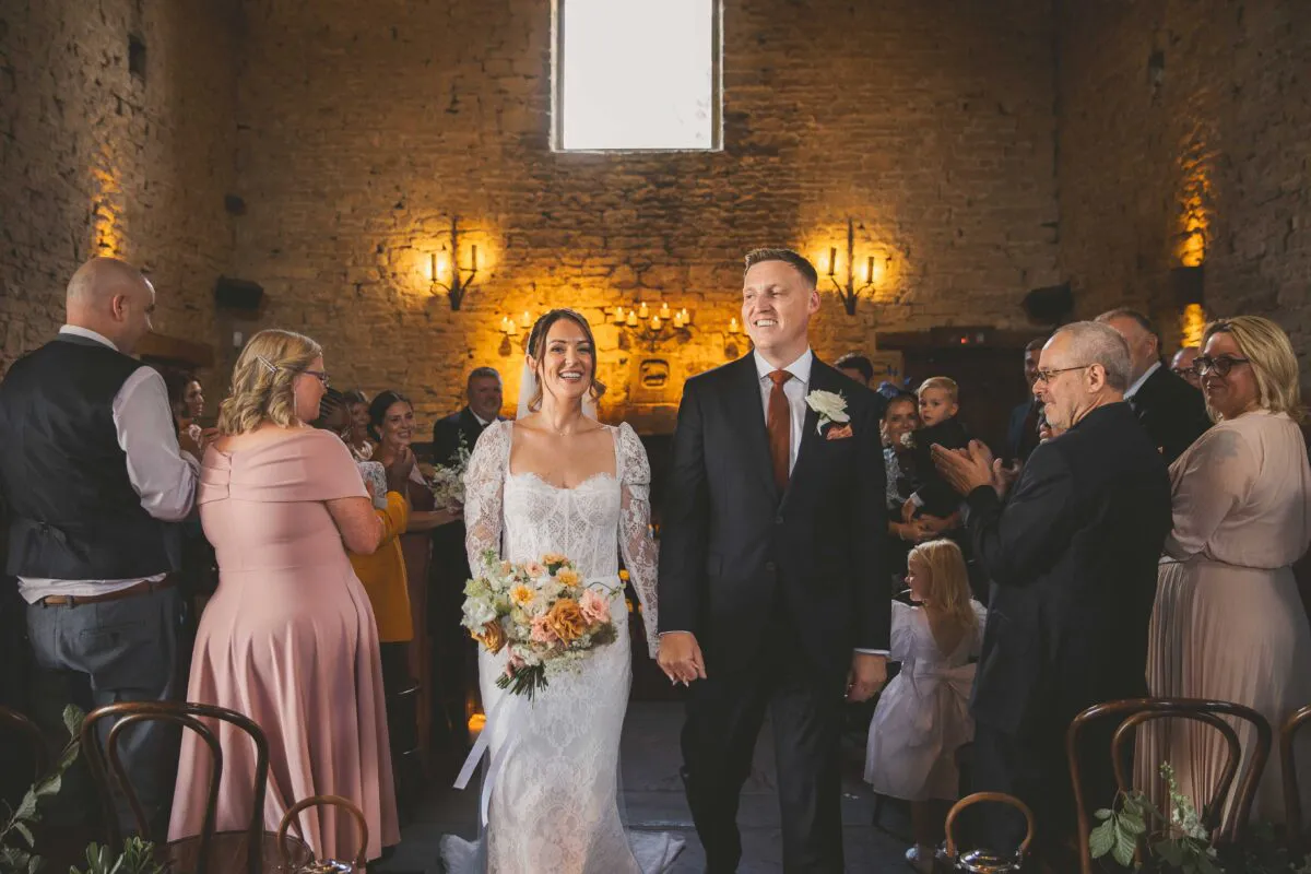 A smiling newlywed couple walks hand in hand down the aisle of a rustic stone barn, surrounded by applauding family and guests.