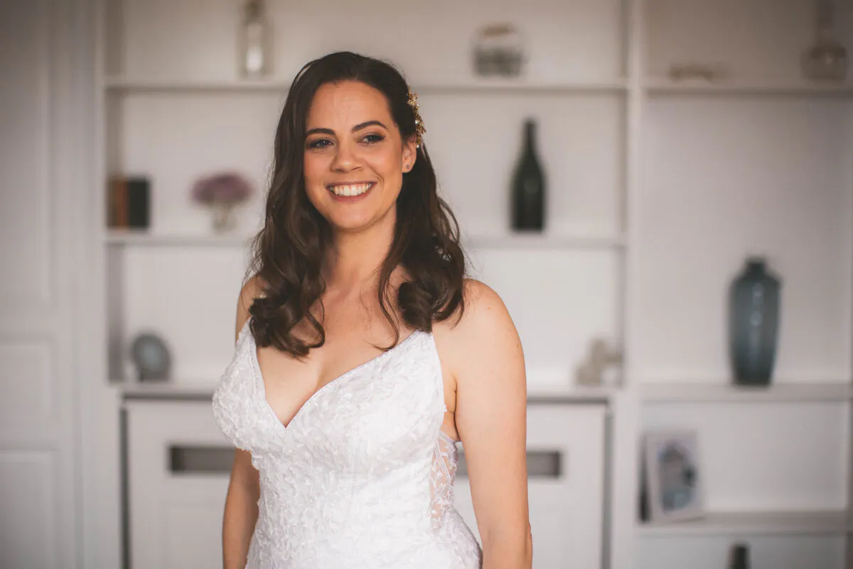 A smiling bride wearing a lace wedding gown with a plunging neckline stands indoors during her morning preparations.