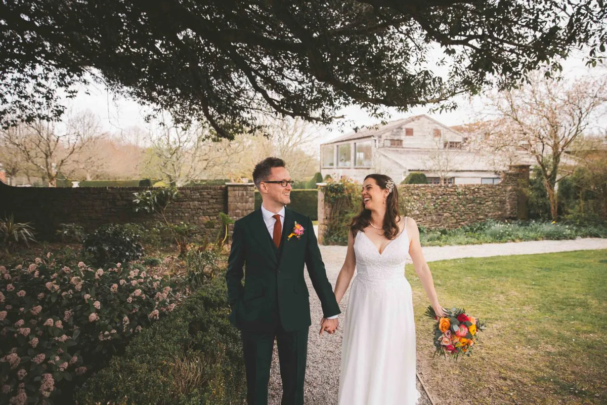 A newlywed couple laugh and hold hands while walking along a gravel path in a manicured garden with a stone barn in the background.