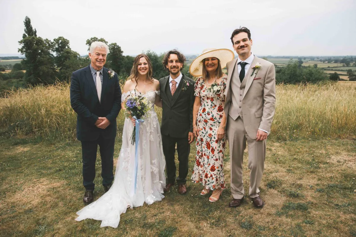 A happy wedding party consisting of a bride holding a wildflower bouquet, a groom, parents, and a guest posing outdoors against a scenic backdrop of rolling green hills under an overcast sky.