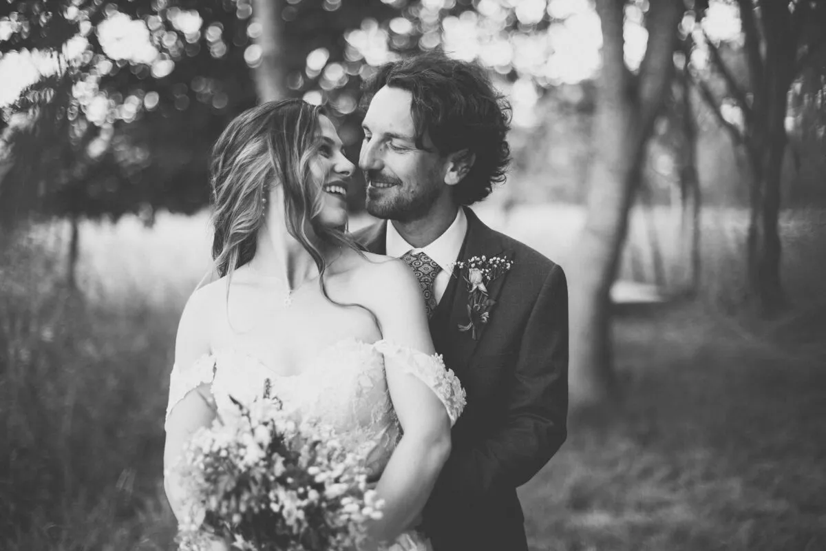 A newlywed couple shares a tender moment smiling at each other in a woodland setting with soft bokeh lighting.