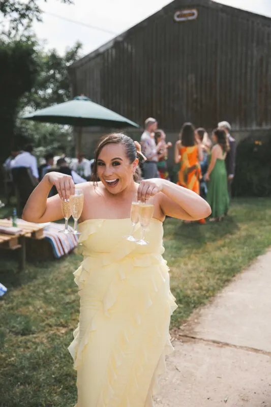 A joyful wedding guest in a yellow ruffled dress playfully holds up four glasses of champagne during an outdoor reception in front of a rustic barn.