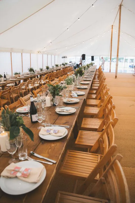 An elegant marquee wedding reception features long wooden banquet tables set with white linens, greenery runners, and warm candlelight underneath festoon lighting.