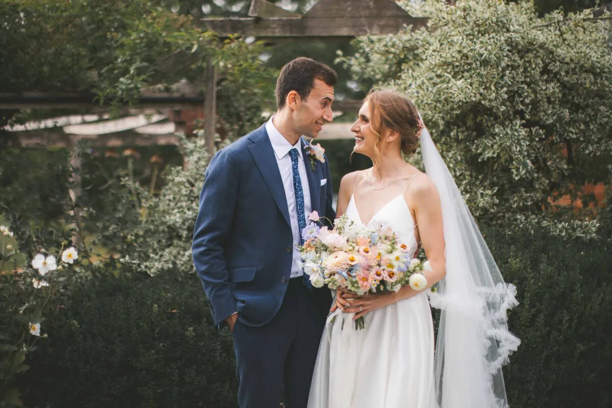 A newlywed couple smiling lovingly at each other outdoors in a lush garden setting, the bride holding a colourful pastel bouquet.