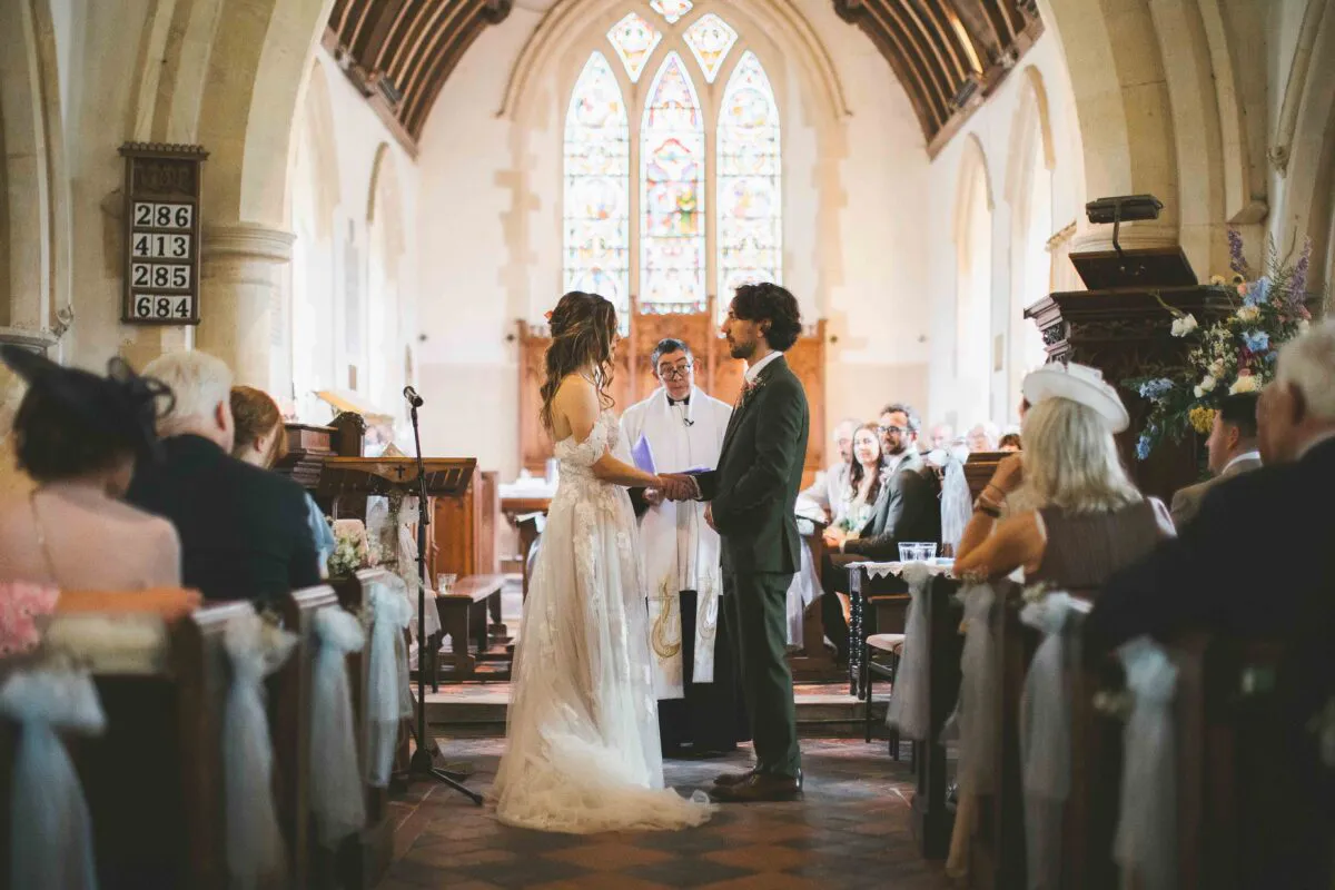 A bride in a floral applique gown and a groom in a green suit stand holding hands at the altar during a church wedding ceremony.