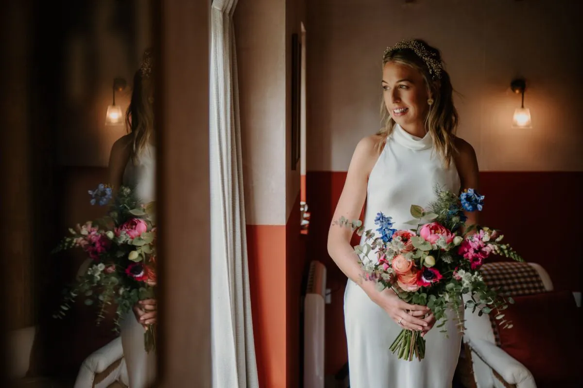 A smiling bride holding a colourful bouquet of roses and delphiniums stands in an interior room with a mirror reflecting her profile.