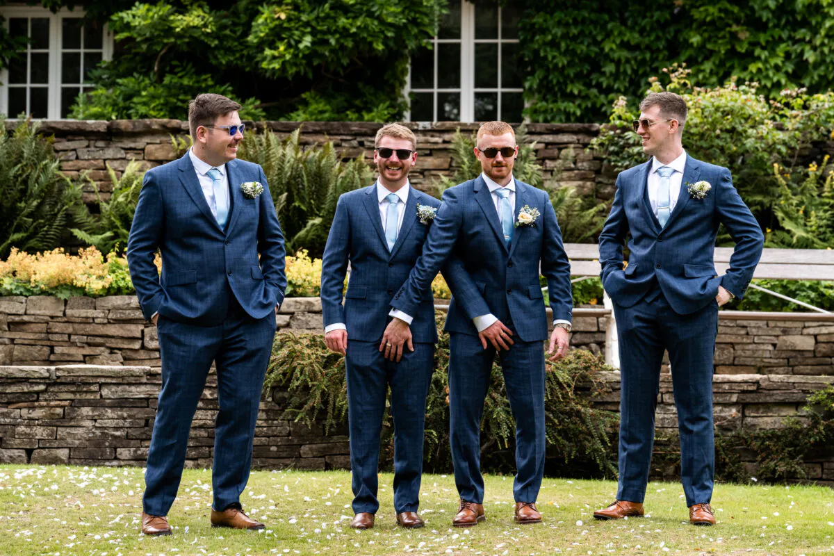A group of four groomsmen wearing matching blue suits and sunglasses share a playful moment laughing together on a manicured lawn.