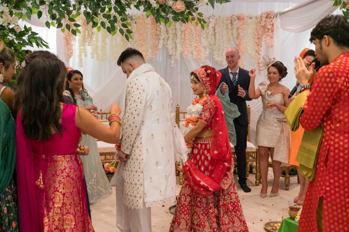 A bride and groom in traditional red and white south asian wedding attire stand beneath a floral archway inside a decorated marquee while guests look on and applaud.