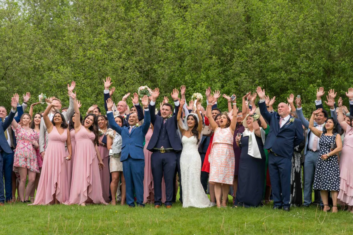 A large, joyful wedding party and guests raise their hands in celebration outdoors against a lush green backdrop on a sunny wedding day.