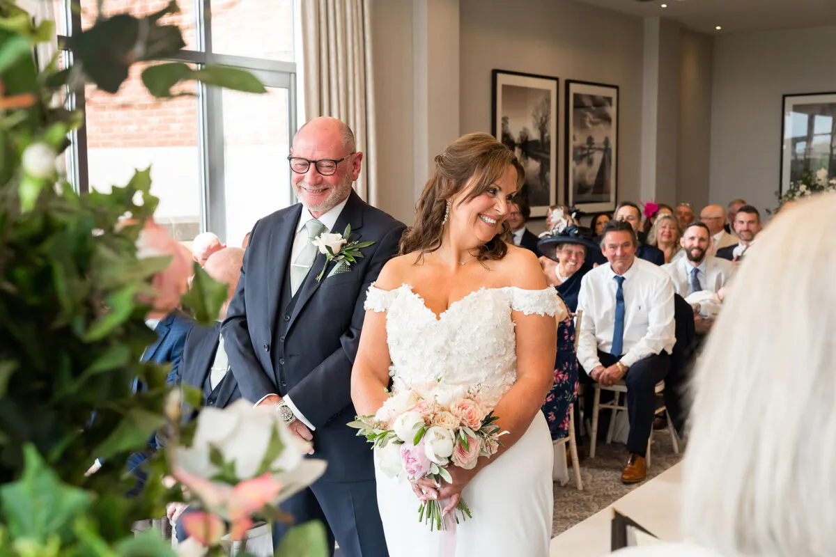 A smiling mature bride holding a floral bouquet stands beside her groom at the front of a light filled ceremony room.