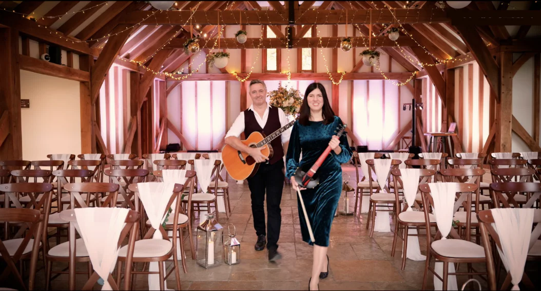 A male acoustic guitarist and a female violinist walk down the aisle of an oak-framed barn venue decorated with twinkling string lights.