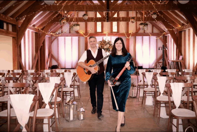 A male acoustic guitarist and a female violinist walk down the aisle of an oak-framed barn venue decorated with twinkling string lights.