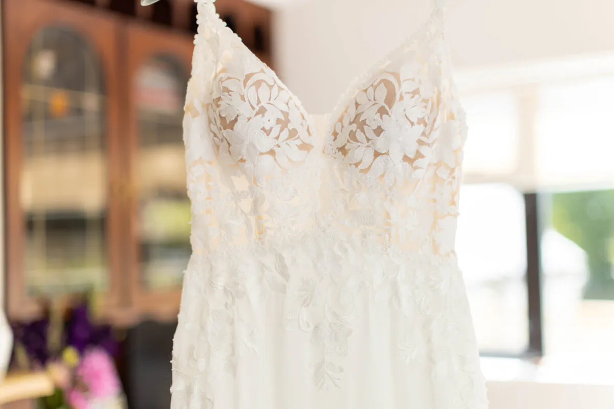 An intricate lace wedding dress hangs in a bright room, showcasing its delicate floral applique and sheer corset bodice.