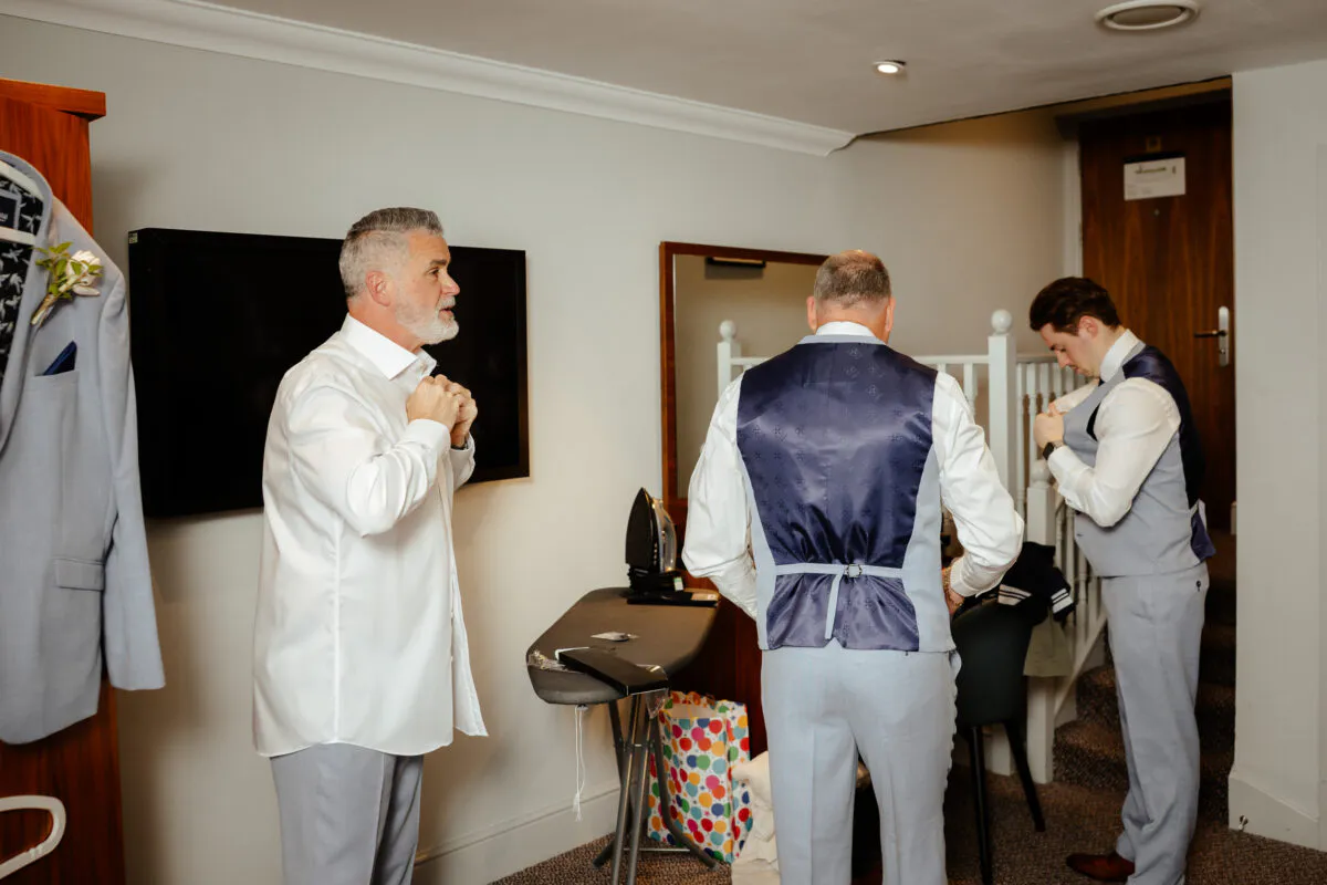 Three men in crisp white shirts and grey trousers prepare for a wedding, with one buttoning his shirt and others adjusting their waistcoats.
