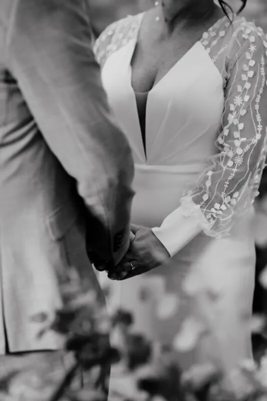 A close up black and white shot of a bride and groom holding hands during their wedding ceremony, highlighting the bride's detailed sheer long sleeves and engagement ring.