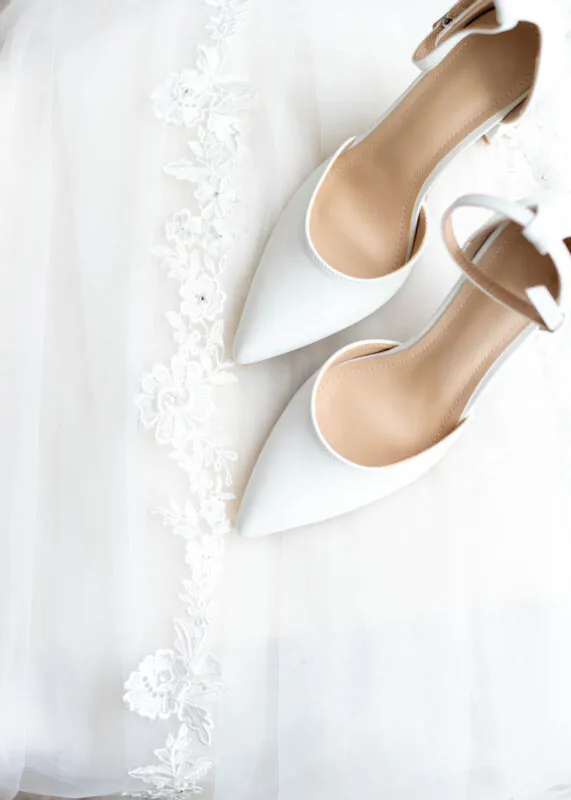 An elegant flat lay showcases white pointed-toe bridal heels resting gently upon a delicate lace-trimmed wedding dress.