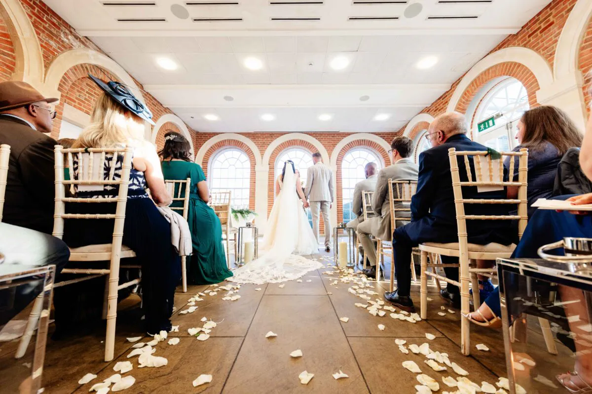 A wide angle view of a civil wedding ceremony inside a light-filled room featuring exposed brick walls and arched windows, with guests seated on chairs facing the bride and groom at the end of a petal-strewn aisle.