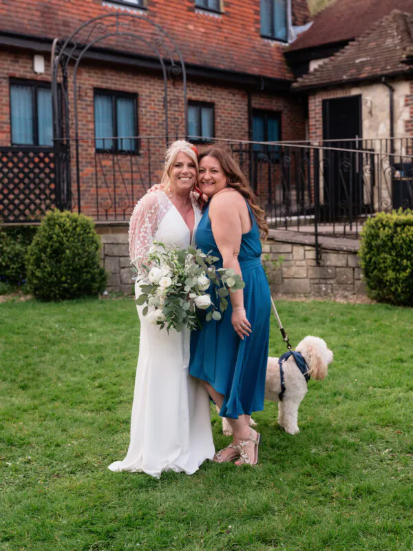 A smiling mature bride in a long-sleeved white crepe gown with sheer embroidered sleeves poses with a guest in a blue dress and a small dog on a lead in a lawn garden setting.