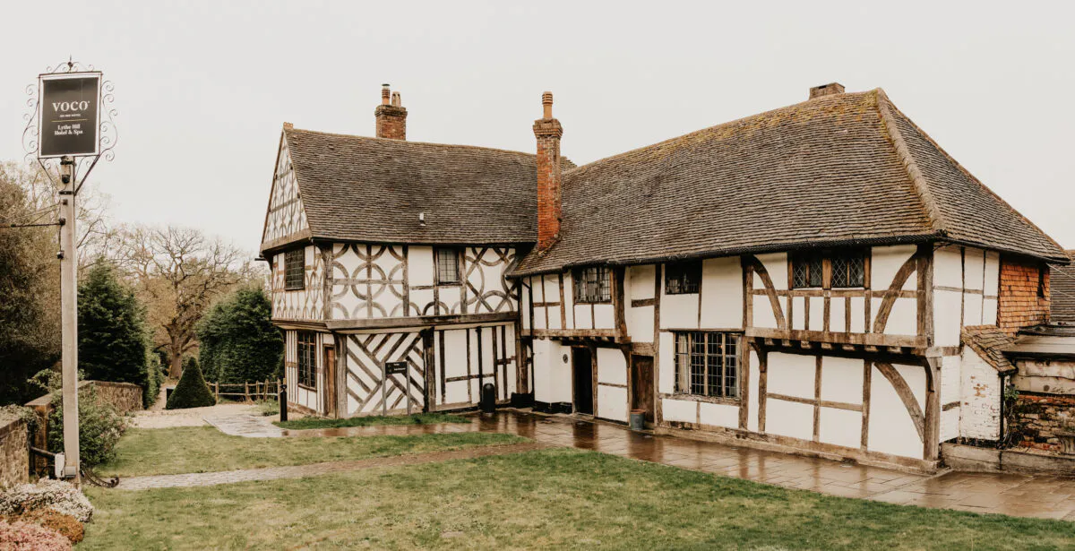 An exterior view of a historic timber-framed Tudor building set against an overcast sky with a paved pathway and lawn in the foreground.
