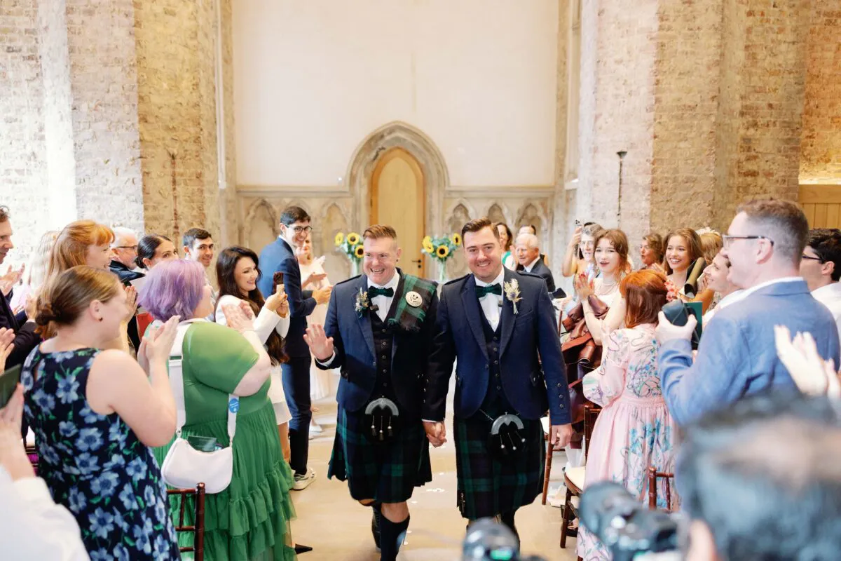 Two smiling grooms in traditional tartan kilts walk hand in hand down the aisle of a historic stone church while cheering guests applaud.