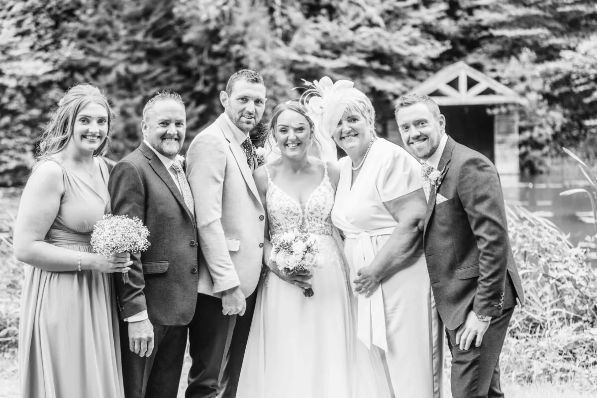 A happy wedding group portrait featuring the bride and groom standing alongside family members and wedding party in an outdoor lakeside setting.