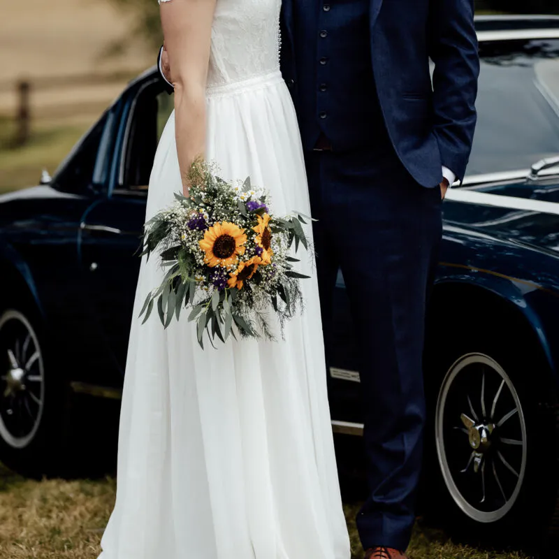 A bride and groom pose by a classic dark car, with the bride holding a vibrant wildflower bouquet featuring sunflowers.
