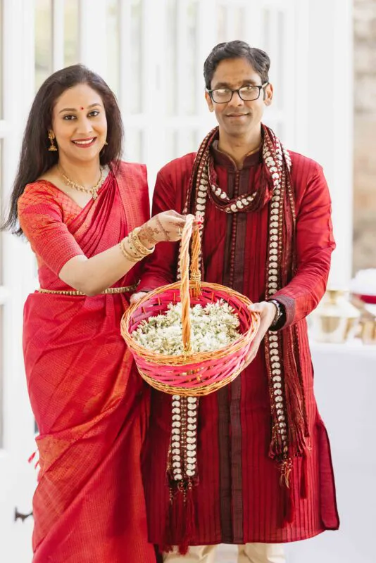 A smiling couple in matching vibrant red traditional attire holds a woven basket filled with white flowers during a wedding celebration.
