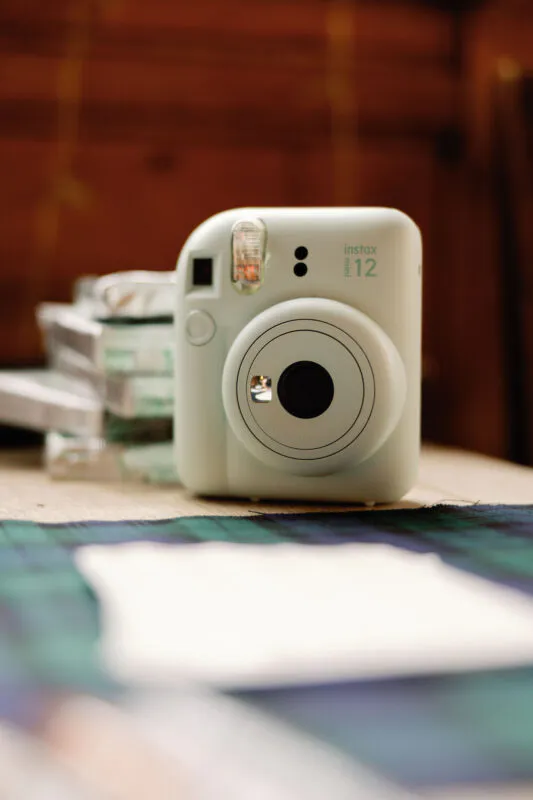 A pastel green instant camera sits on a wooden surface beside a tartan fabric and a blurred white card.