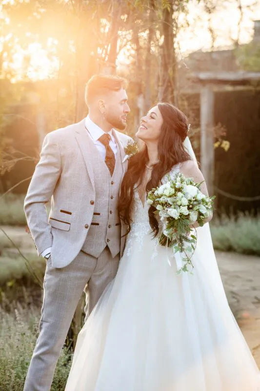 A newlywed couple exchange a loving glance outdoors in a sunlit garden during golden hour, with the bride holding an elegant white floral bouquet.