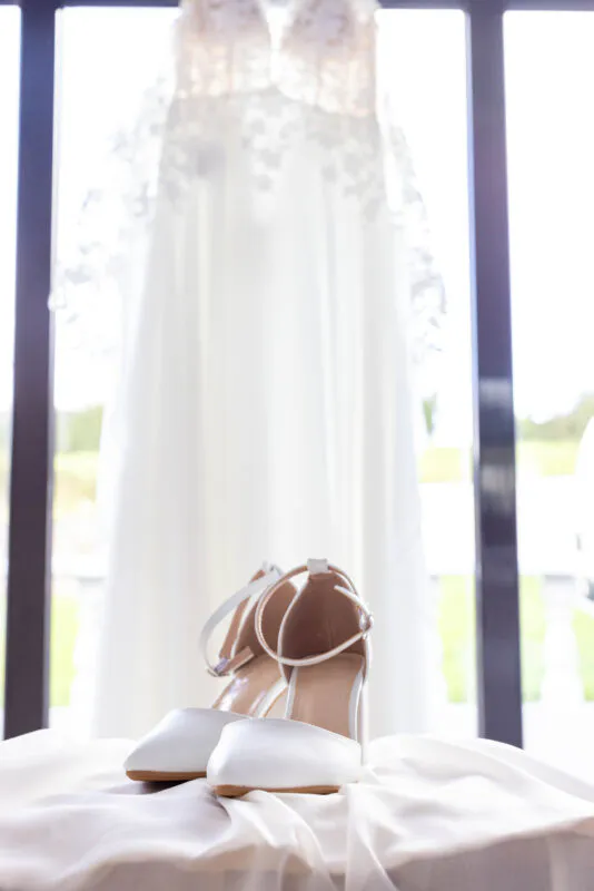 A pair of white block-heel bridal shoes rests on fabric in the foreground, with a lace wedding dress hanging in the brightly lit doorway behind.