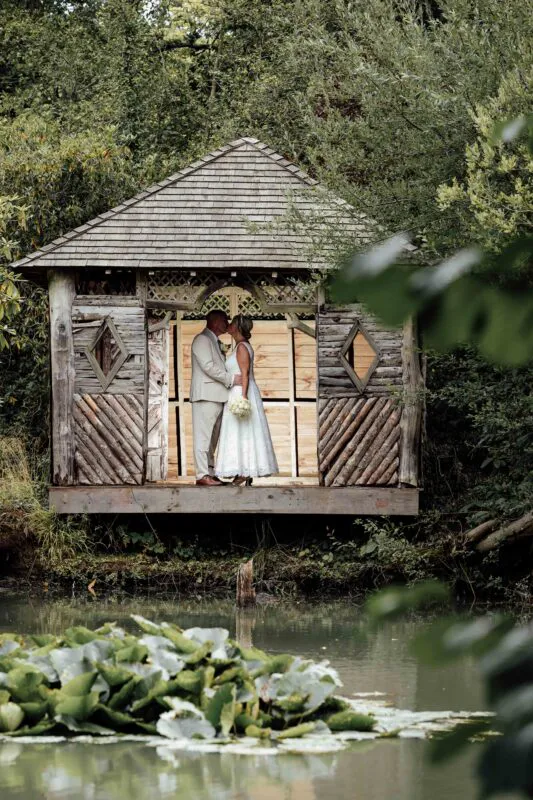 A newlywed couple shares a tender kiss inside a rustic wooden summer house set beside a tranquil lake surrounded by lush green woodland foliage.