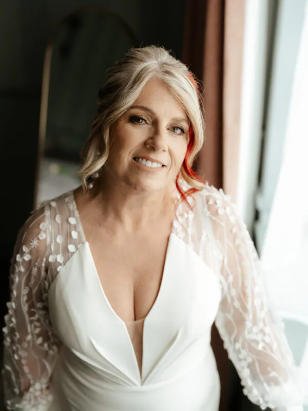 A mature bride with blonde and red hair smiles gently near a window, wearing a V-neck wedding dress with sheer floral applique sleeves.