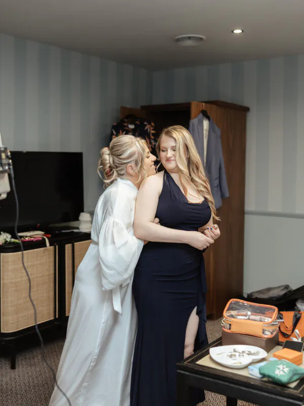 Two smiling brides share a tender embrace indoors during bridal preparations, with one wearing a white robe and the other in an elegant navy gown.