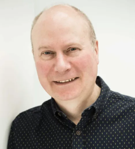 A close-up portrait of a smiling male guest wearing a dark patterned shirt against a bright background.