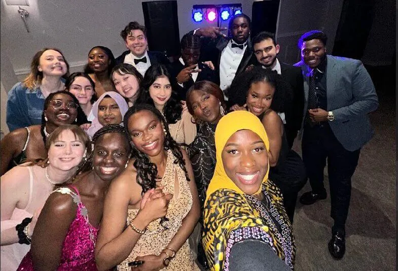 A joyful wedding crowd gathers close together for a group selfie on the dance floor under colourful disco lights.