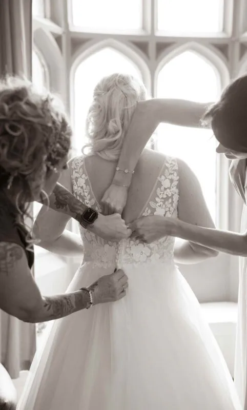 Two bridesmaids help the bride fasten her lace wedding dress in front of a bright window during bridal preparations.