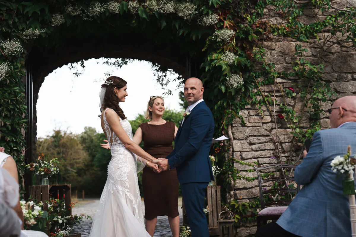A bride in a lace wedding dress and a groom in a navy suit hold hands during an outdoor ceremony beneath a stone archway draped in ivy.