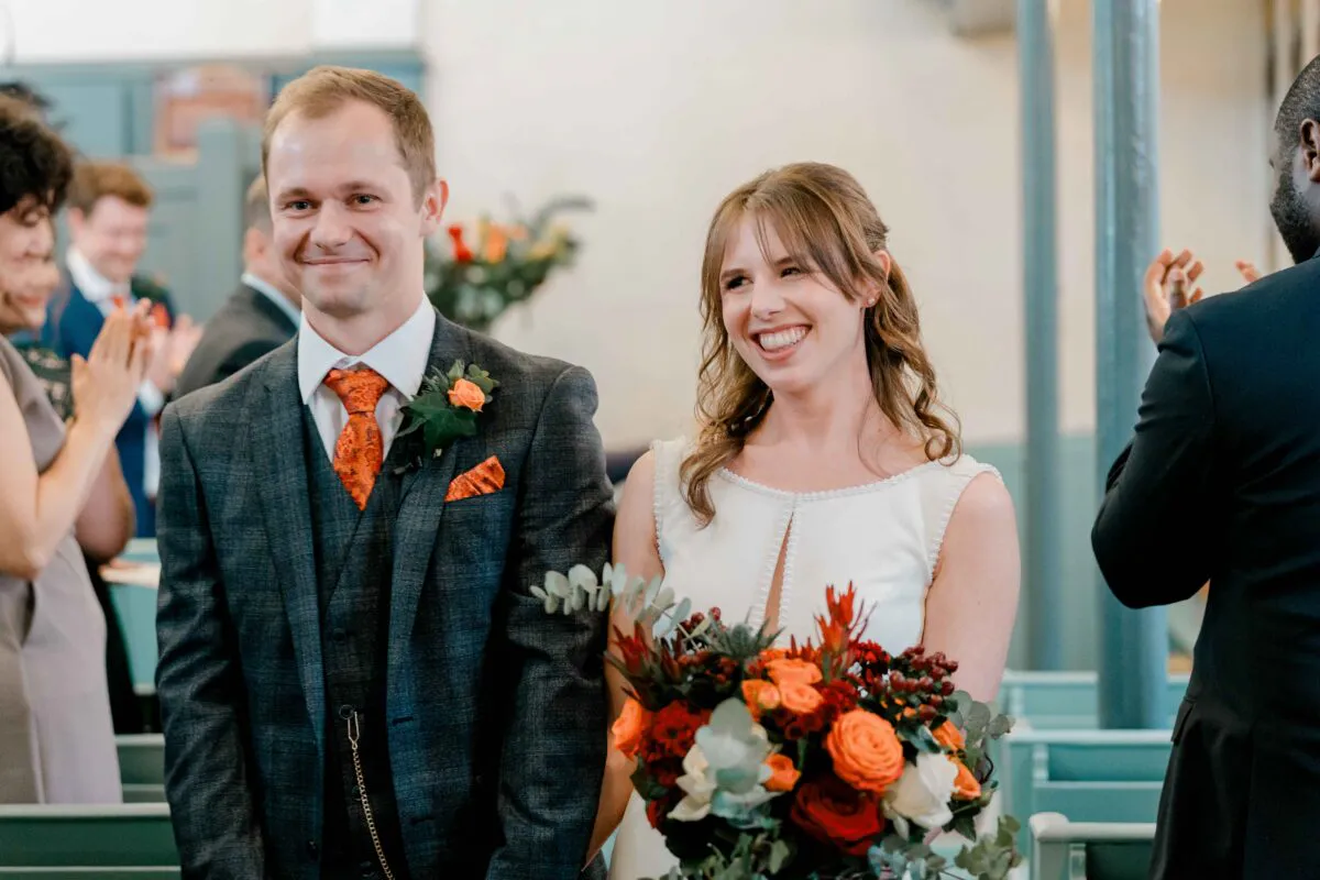 A happily smiling bride and groom stand together holding a vibrant autumn-toned bouquet while guests applaud them during the wedding ceremony.