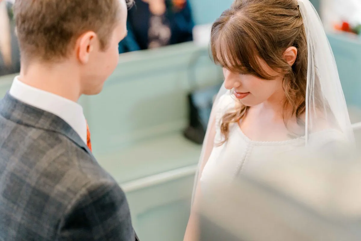 The groom looks lovingly down at his smiling bride with a fringe and veil during their intimate church ceremony.